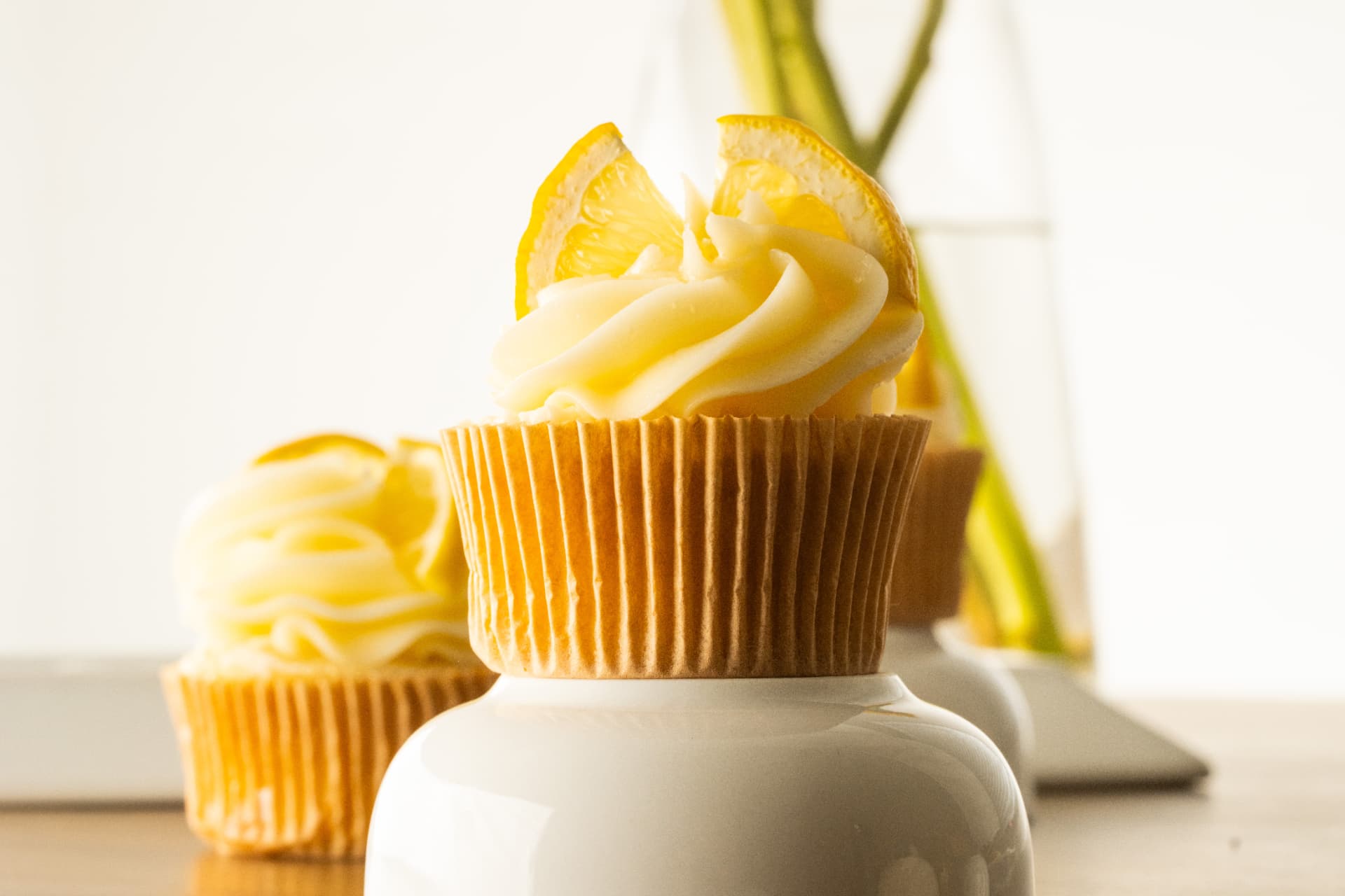 Lemon cupcake on a white ceramic riser with more cupcakes and greenery behind it