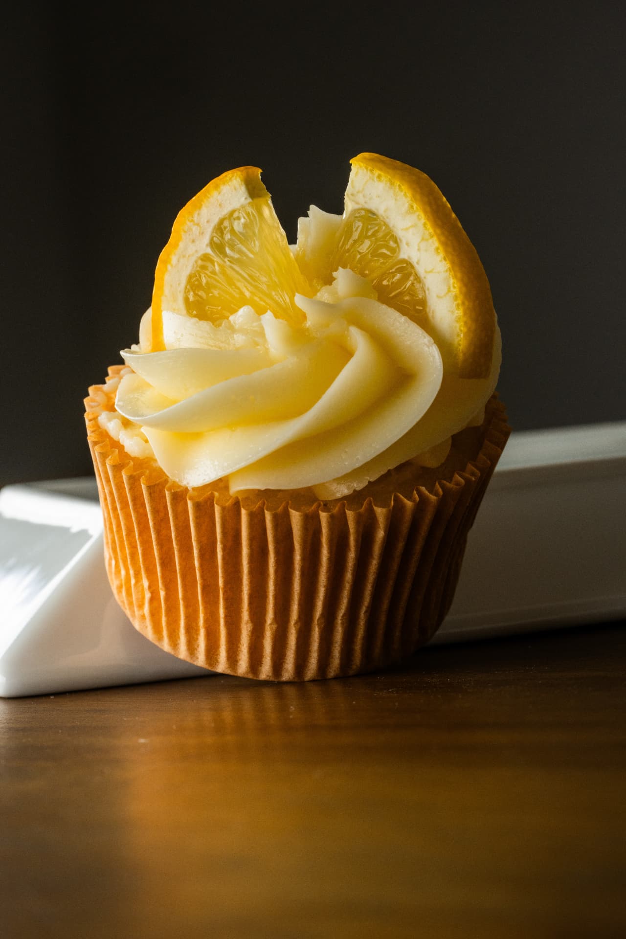Single lemon cupcake with buttercream, white chocolate curls, and a lemon wedge against a dark background