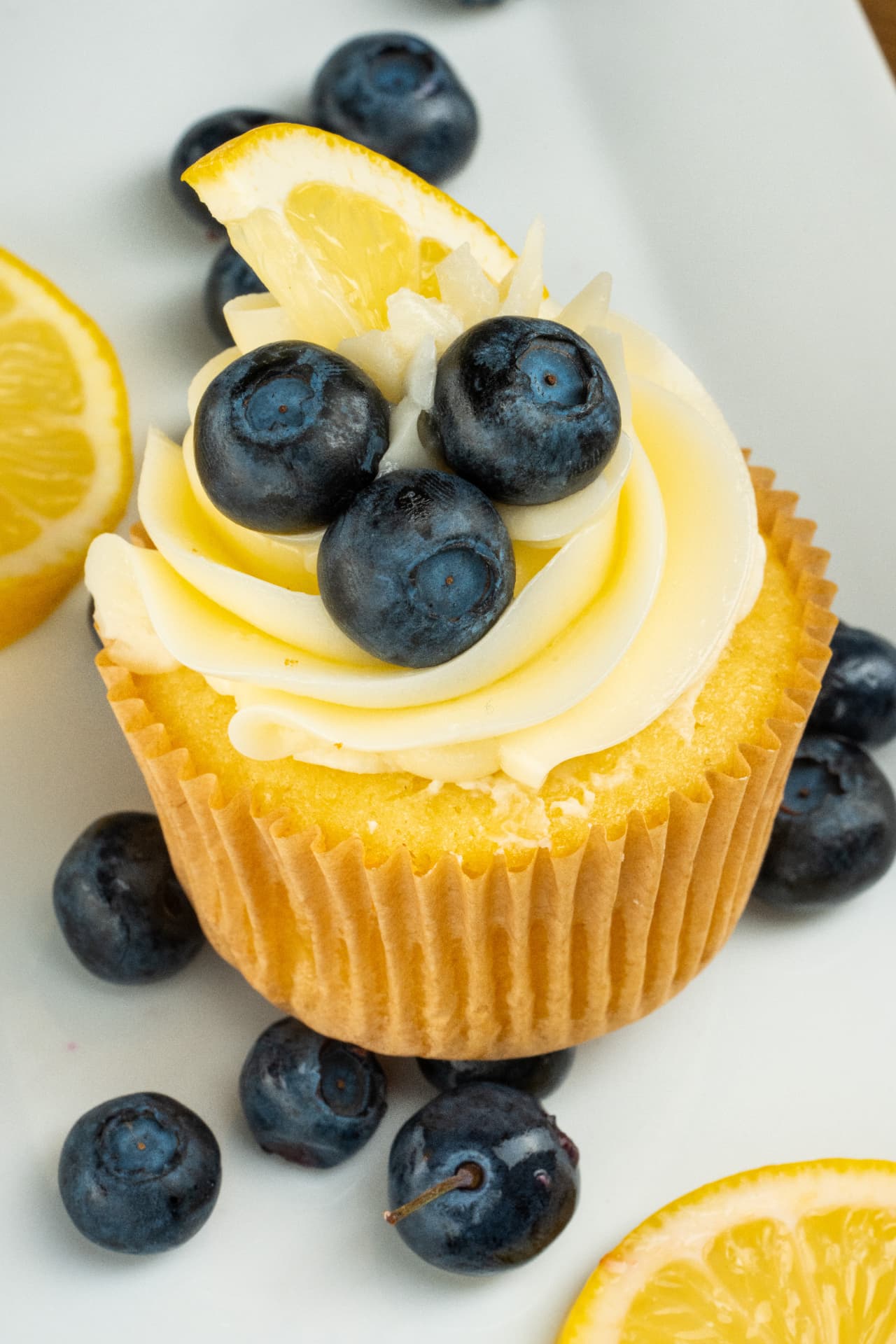 Close-up of a lemon cupcake topped with three blueberries and a lemon wedge on a white surface